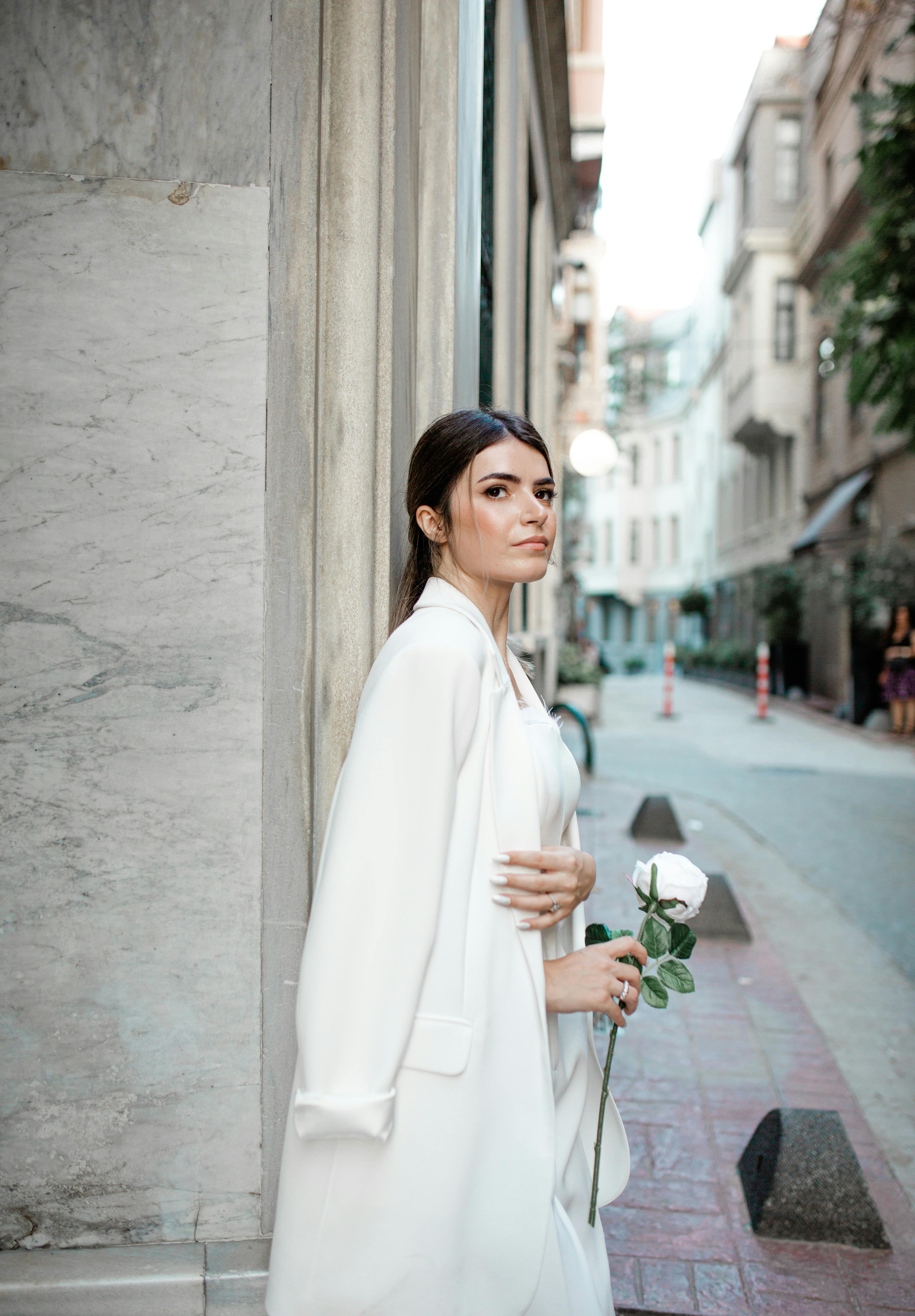 Elegant young woman in white holding a rose against a city street backdrop.