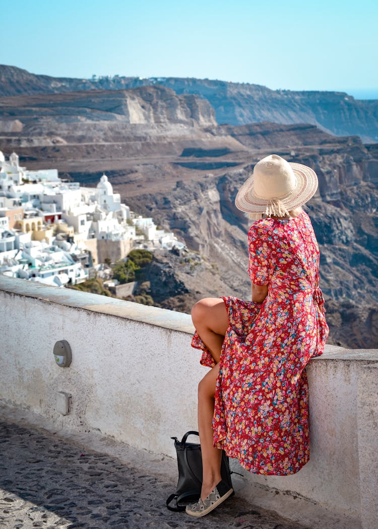 Young Woman Sitting On A Wall And Looking At A View Of A Greek Town And Mountains 