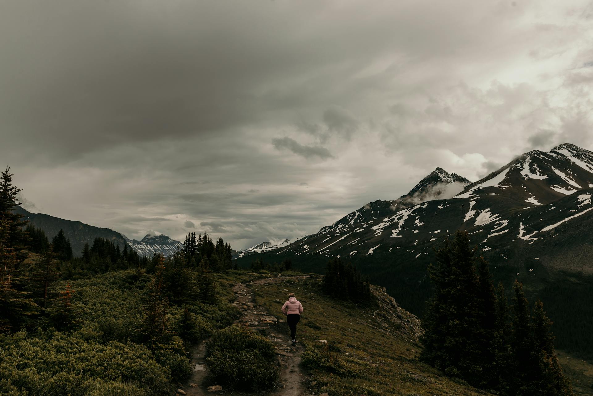 A woman hikes along a rugged trail through a scenic snowcapped mountain landscape.