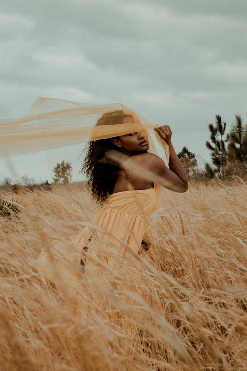 Free An artistic portrait of a woman in a flowing dress standing in a windy wheat field, captured with a dreamy, expressive vibe. Stock Photo
