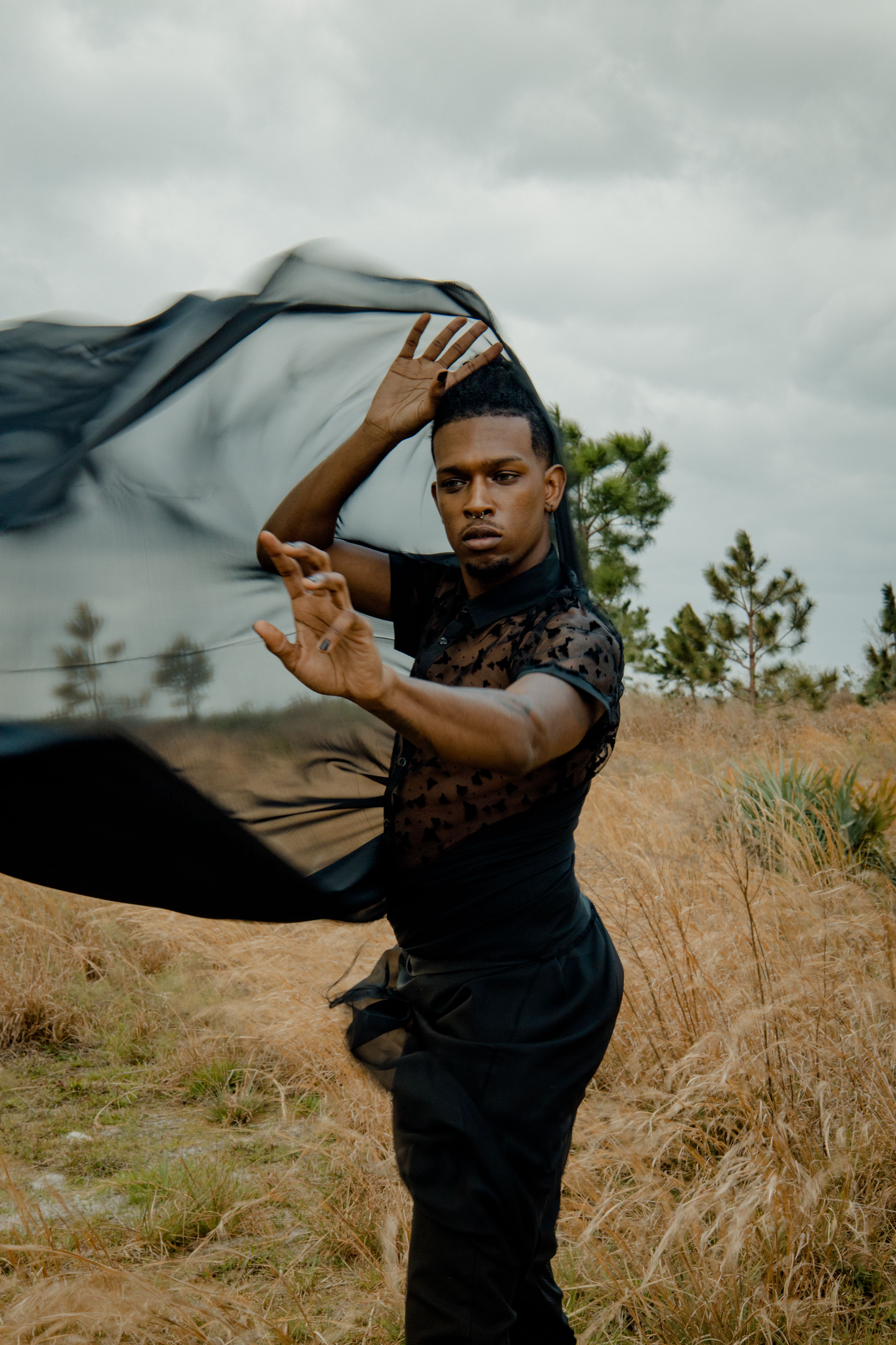 A man elegantly poses with a black cloth in a windy outdoor setting, expressing movement and style.