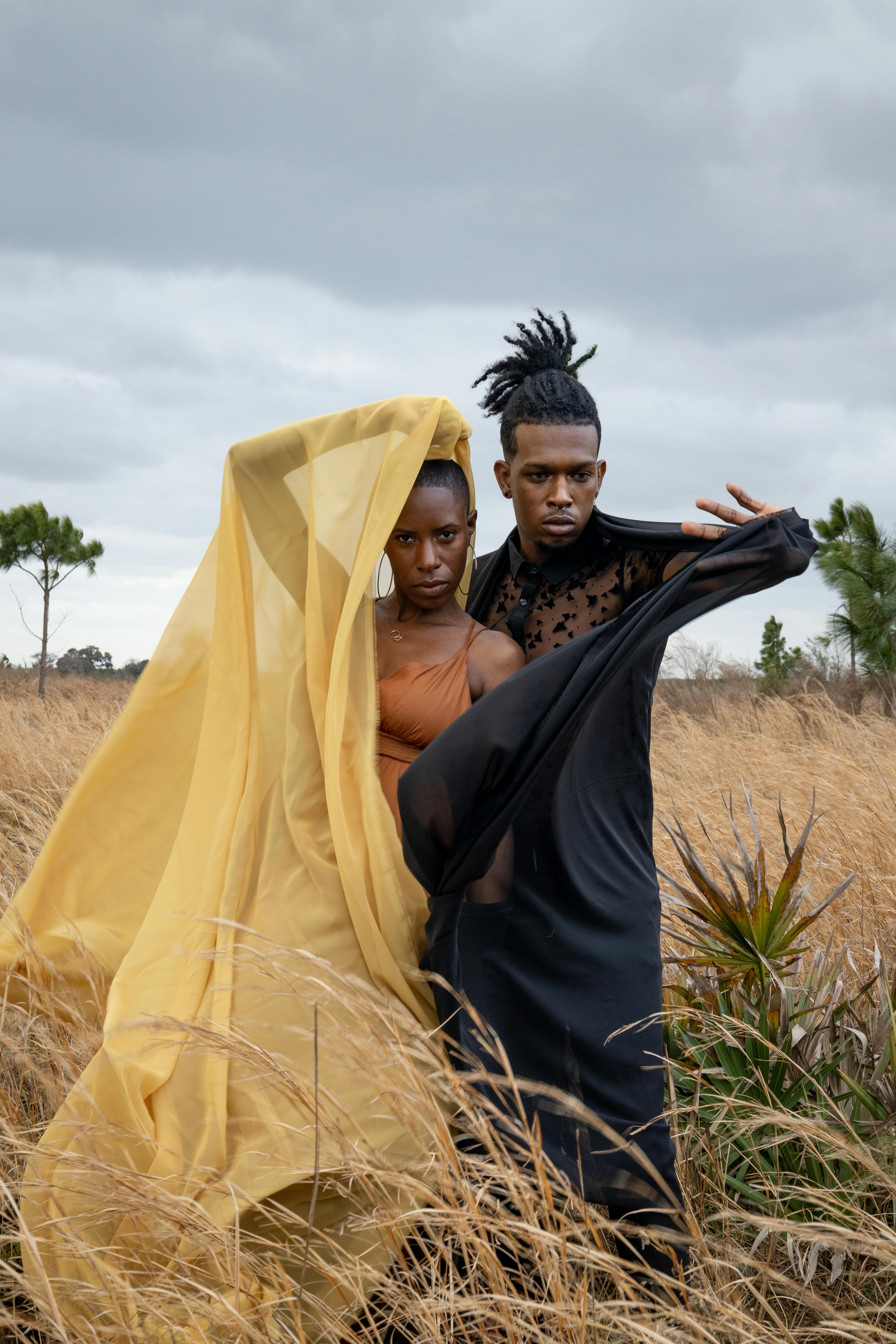 Two models pose dramatically in a field, enveloped in flowing fabrics.