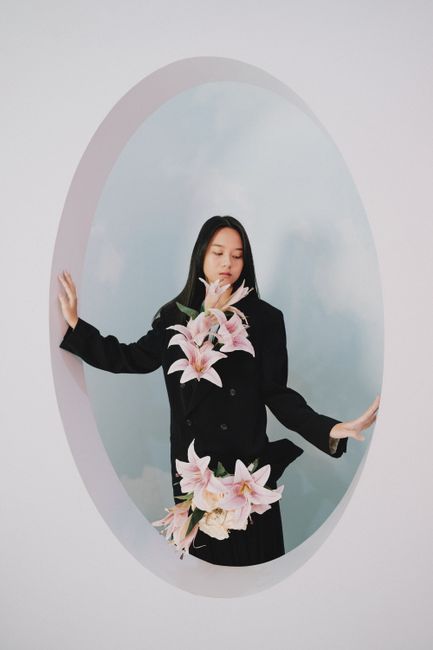 Fashion shoot of a woman in black coat adorned with lilies, eyes closed, against a pastel backdrop.