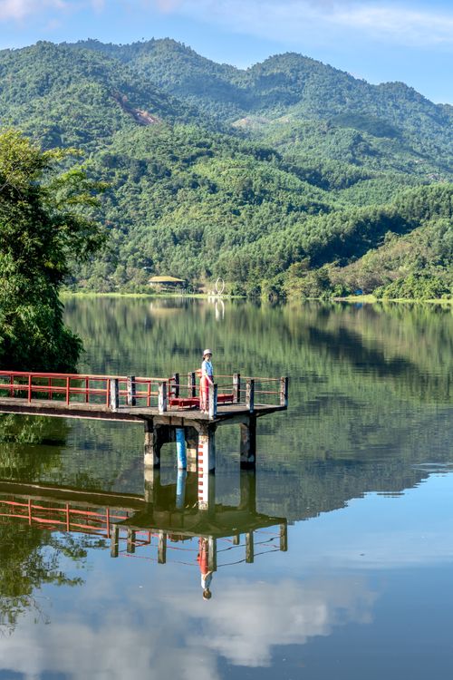 Free Tranquil scene of a lone person on a pier overlooking a beautiful lake surrounded by lush green hills. Stock Photo