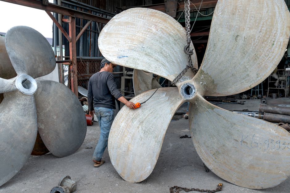 Worker handling large metal ship propellers in an industrial shipyard setting.