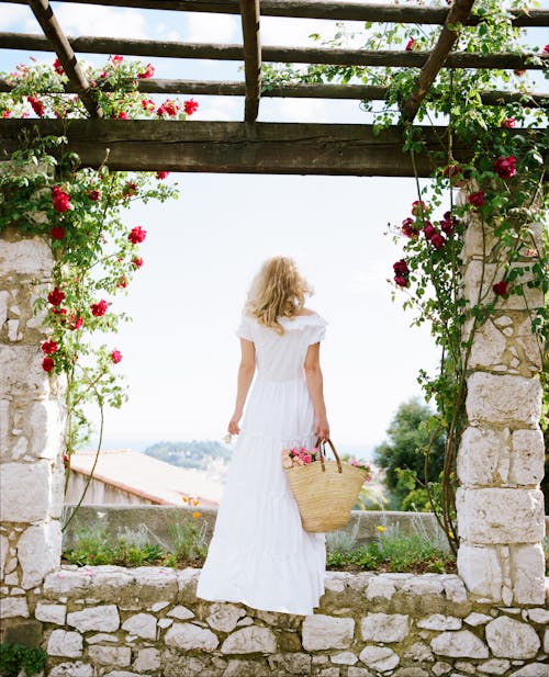 Back View of a Woman in a White Dress Holding a Basket Standing under a Pergola with Roses