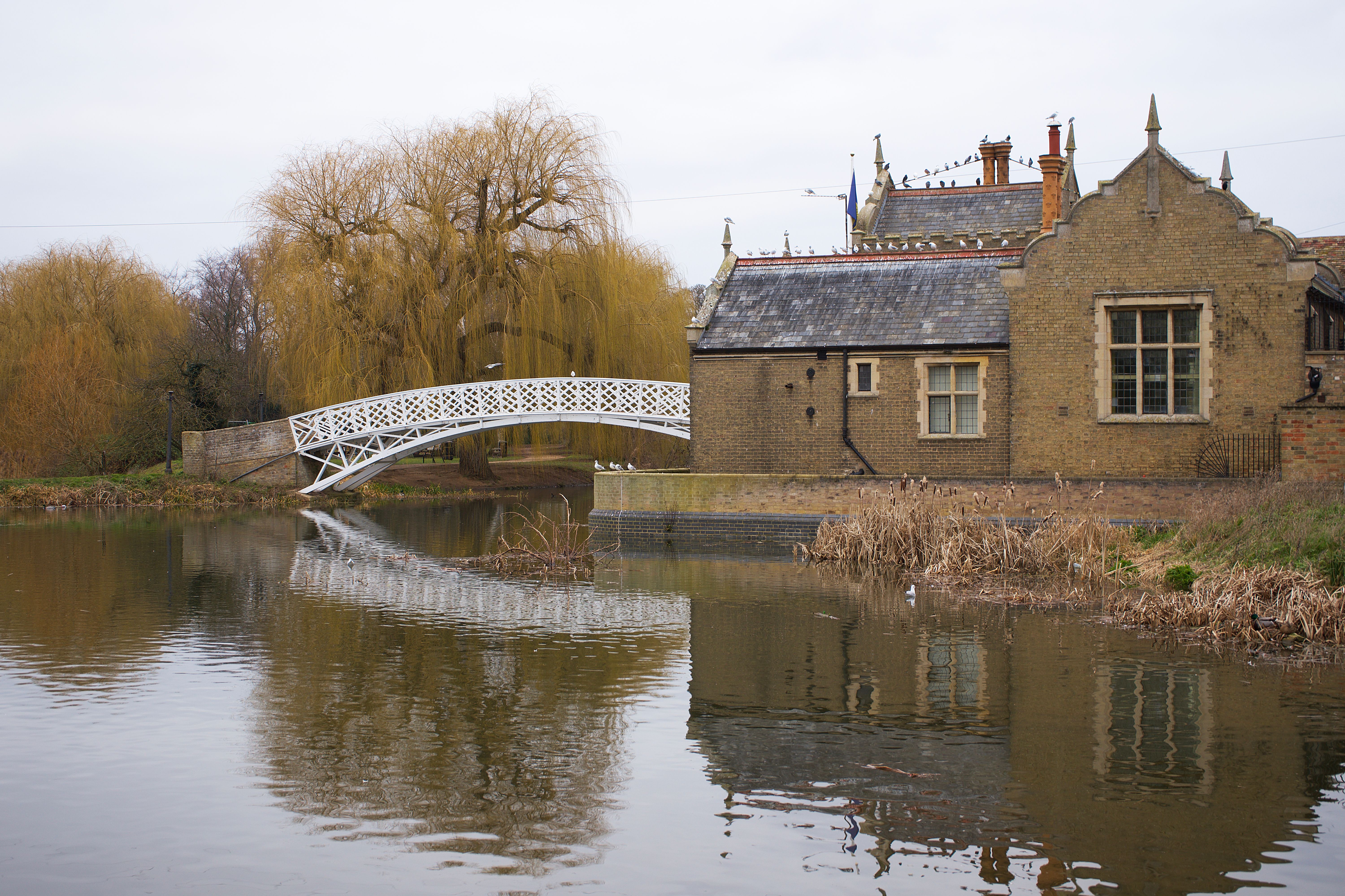 Charming autumn scene of a white bridge and historic house reflecting in a tranquil stream in Godmanchester, UK.
