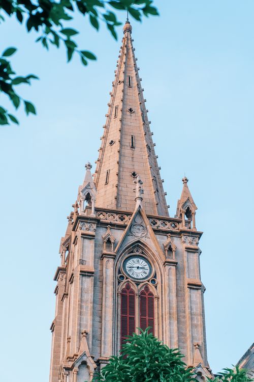 Free A towering Gothic-style church spire reaching for the blue sky, framed by greenery. Stock Photo