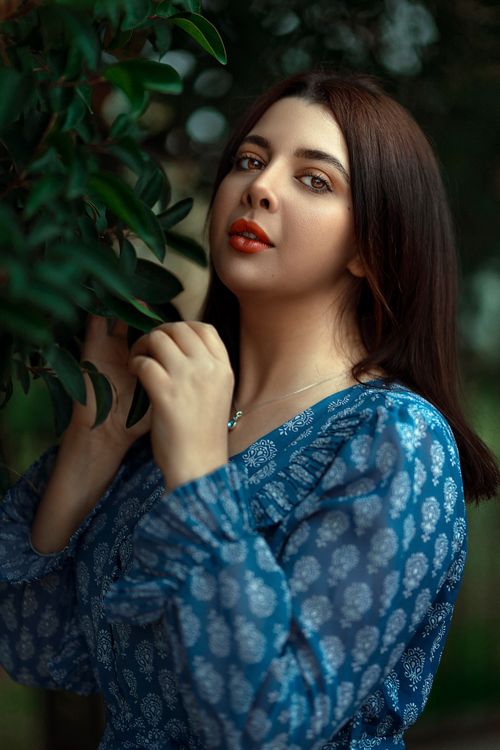 Free Portrait of a young brunette woman in a patterned blue dress posing near green foliage outdoors. Stock Photo