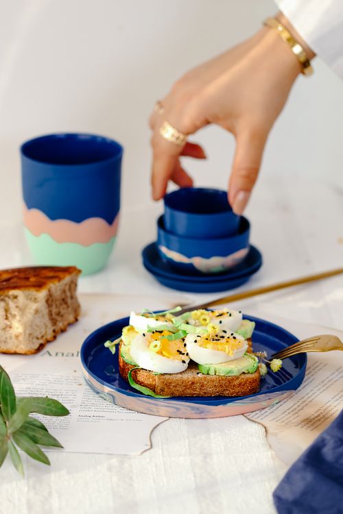 Free Breakfast scene with avocado toast, boiled eggs, and a hand poised to pick up a blue cup. Stock Photo