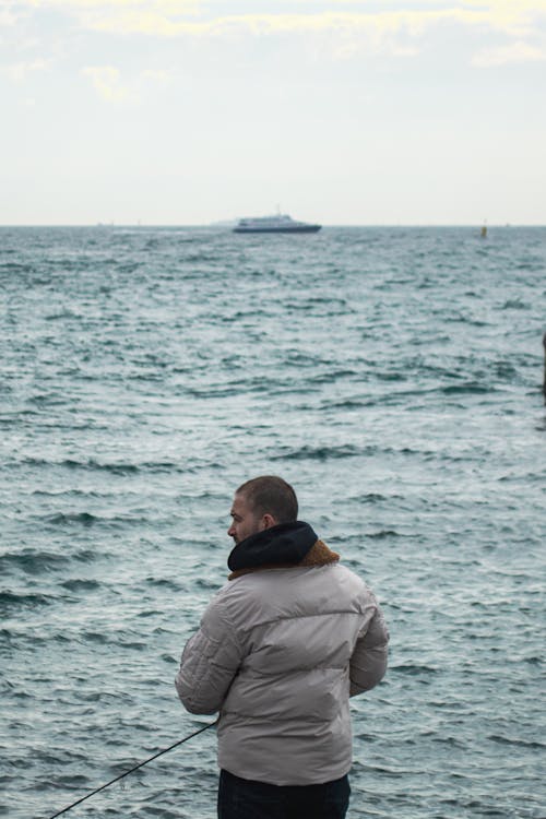 Back View of a Man in a Jacket Standing on the Shore with a Fishing Rod 