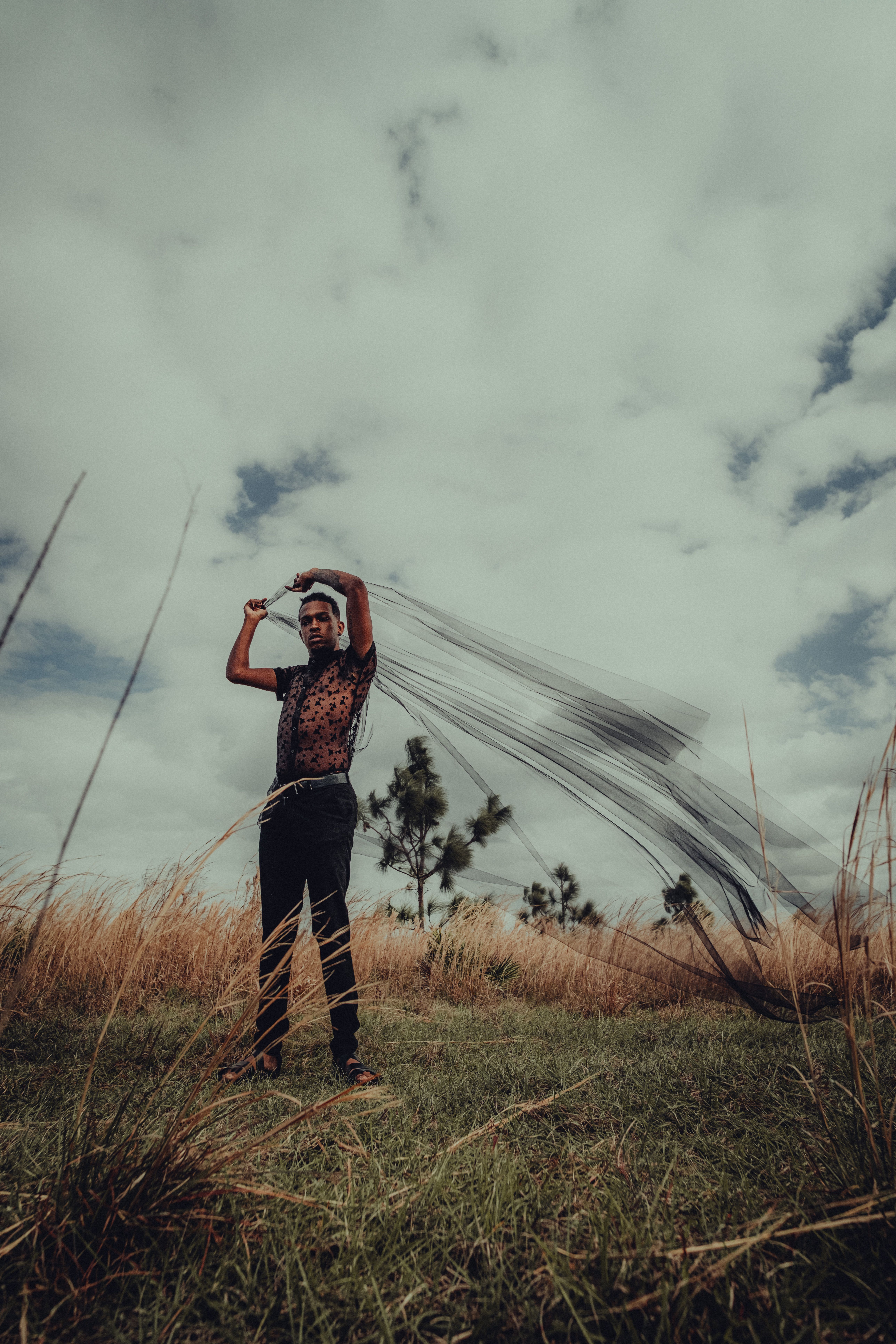 A man poses dramatically with a cloth in a grassy field under a cloudy sky.