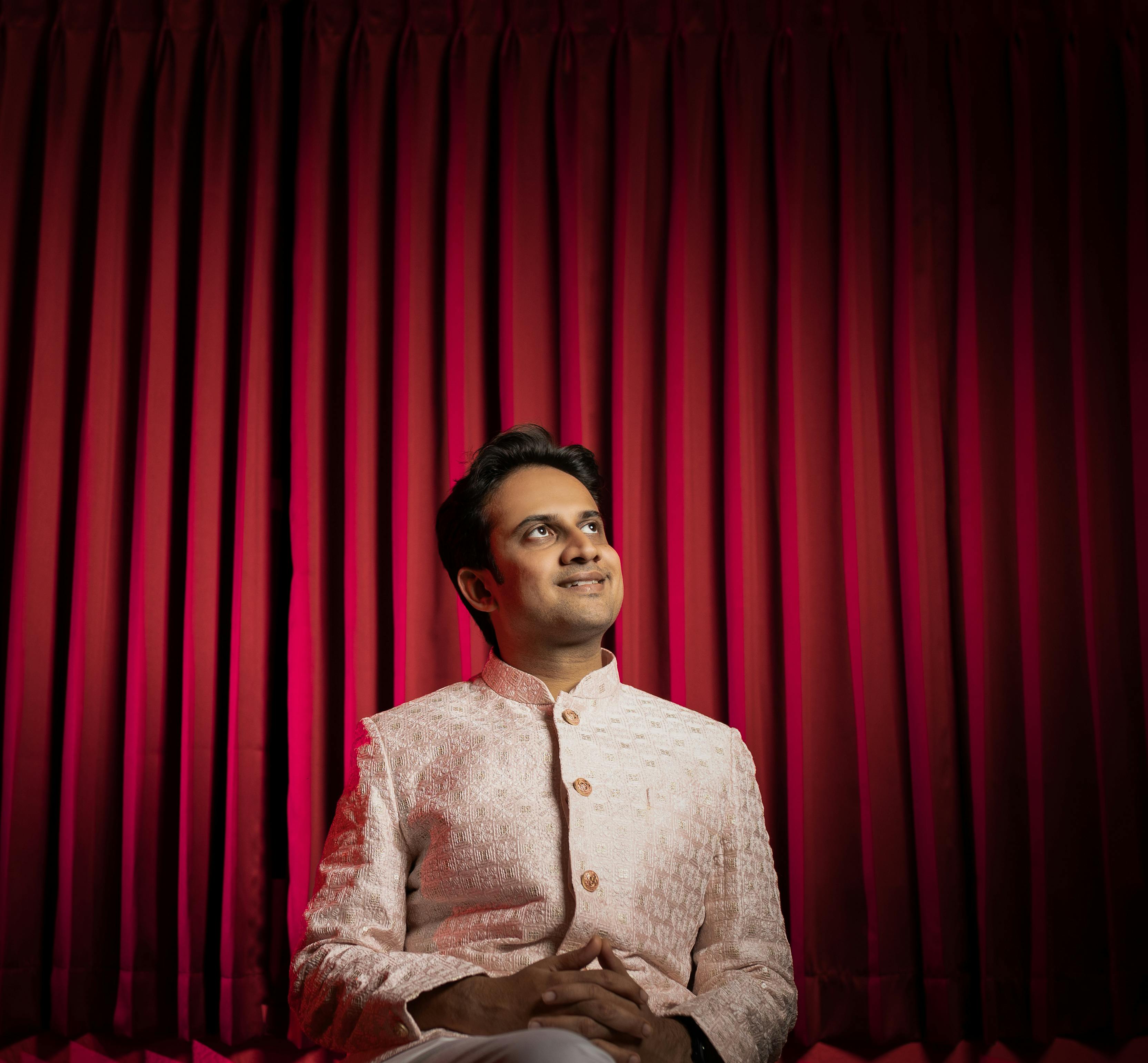A man in formal attire smiles thoughtfully against a vibrant red curtain backdrop.