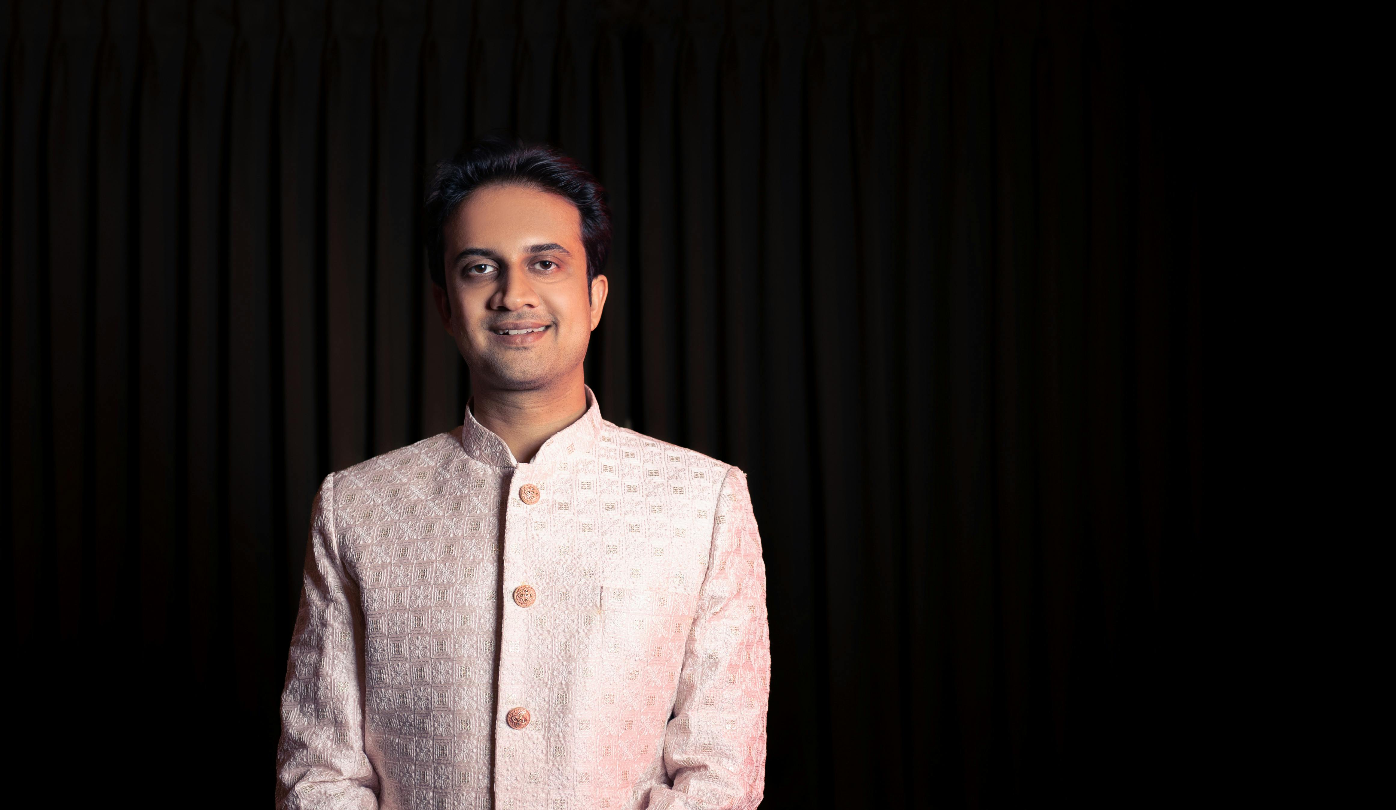 A man in traditional Indian attire posing with a smile against a dark background.