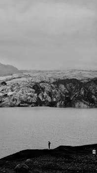 Black and white photo of a lone figure standing by a glacier edge, offering dramatic scenery.