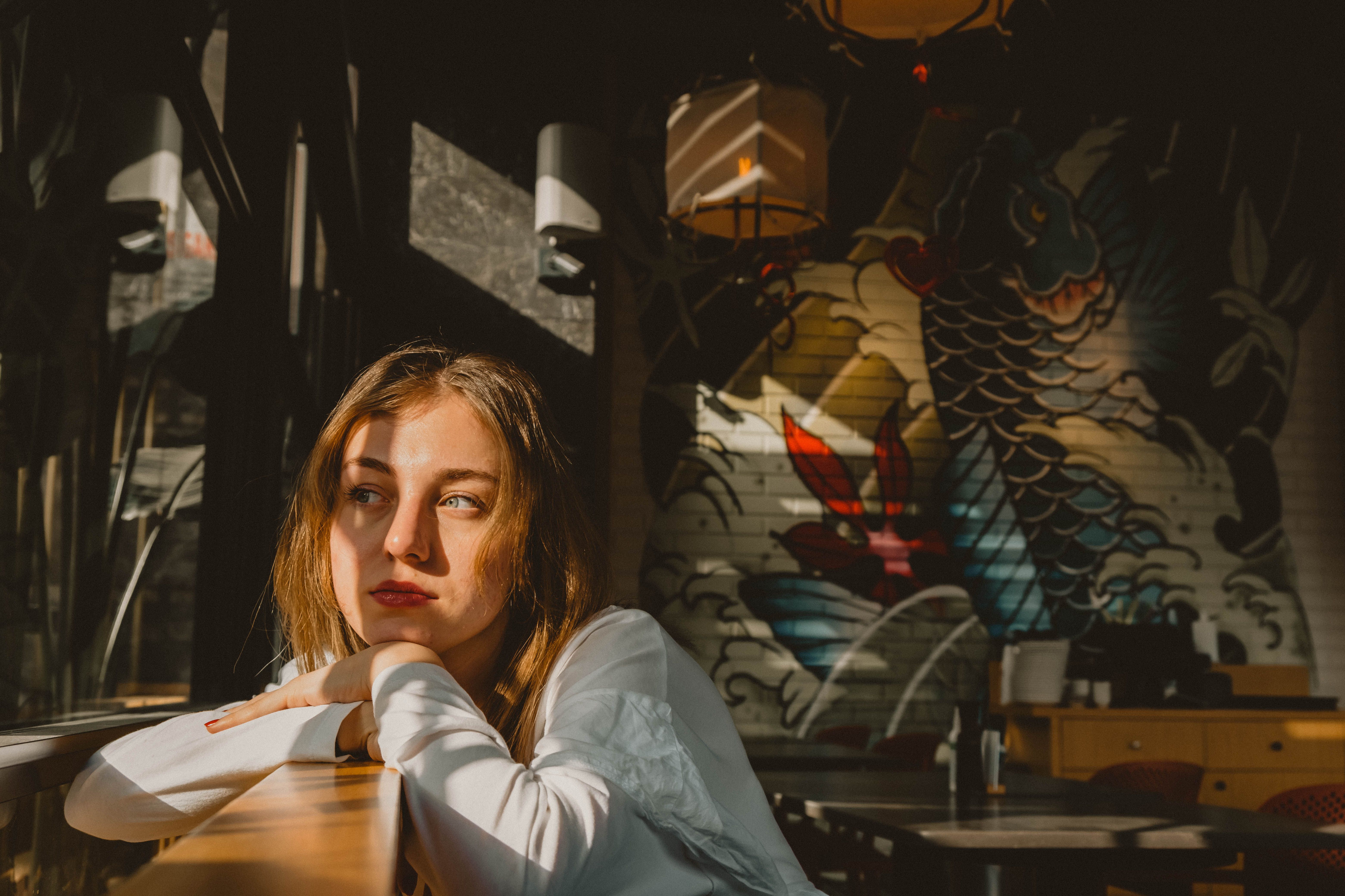 A thoughtful woman gazes outside in a sunlit room with vibrant wall art and soft lighting.