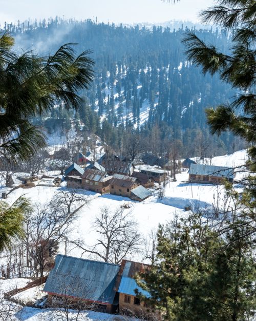 Free Charming village nestled in snow-covered hills under a clear winter sky, surrounded by pine trees. Stock Photo