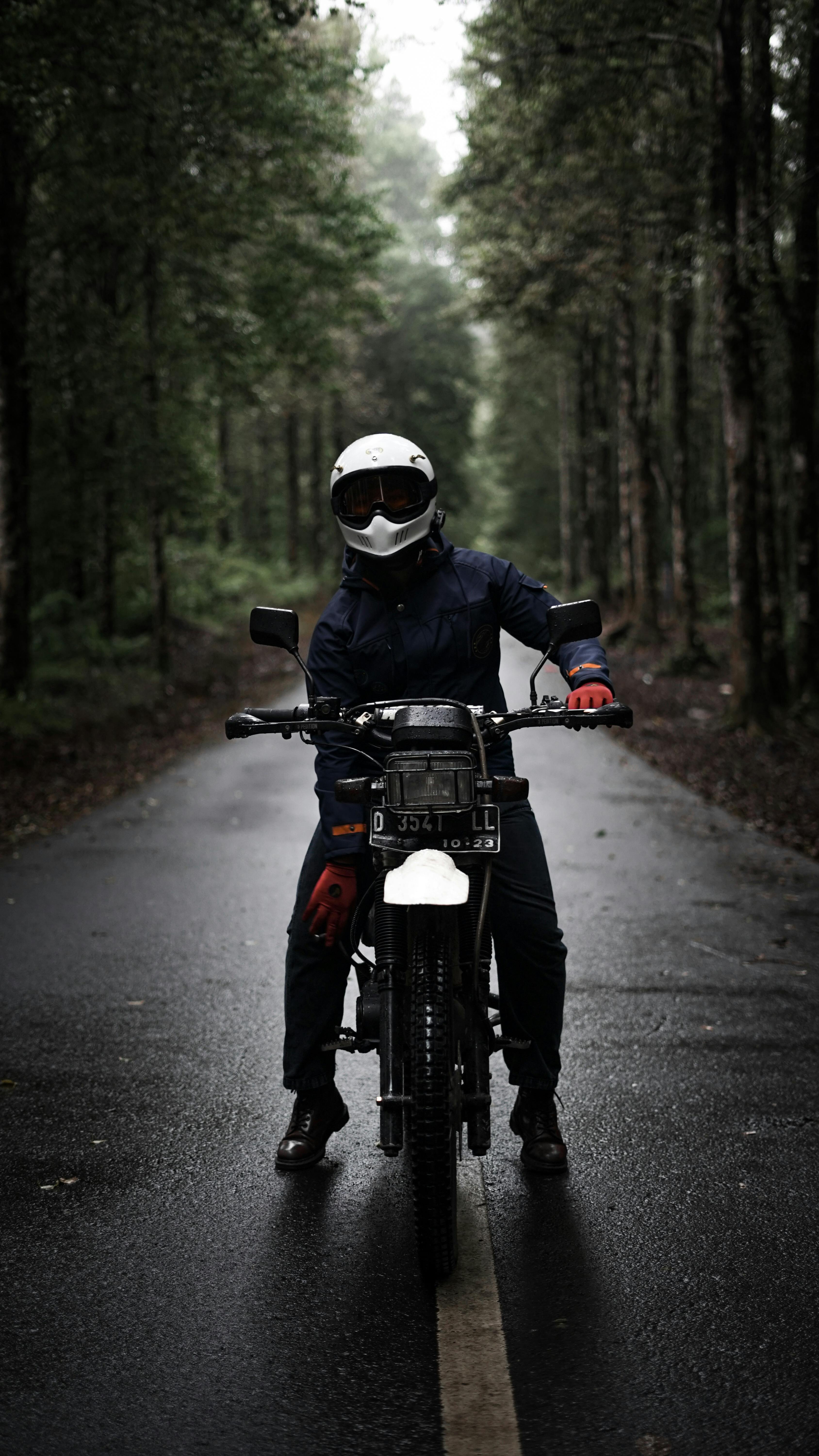 A biker wearing a helmet rides a motorcycle on a wet road through a forest, emphasizing adventure and safety.