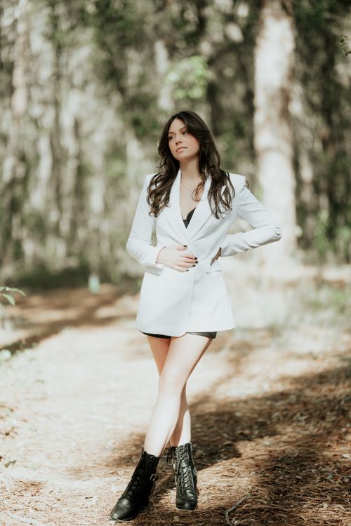 Free Stylish woman posing in a white jacket and black boots in a sunlit forest path. Stock Photo