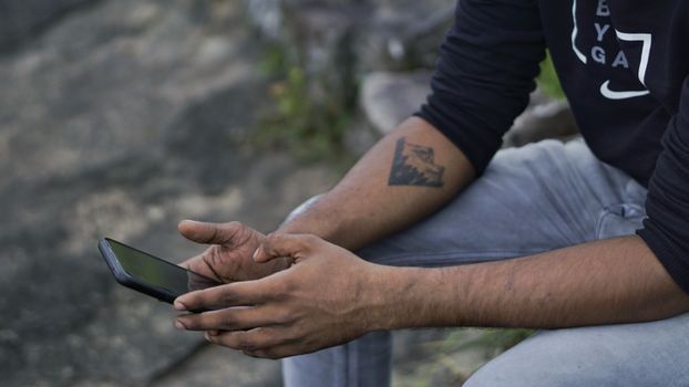 Close-up of a man with an arm tattoo using a smartphone while sitting outside.
