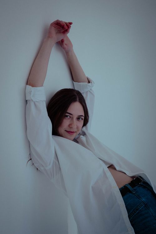 Free Woman posing fashionably in white shirt against a minimalist white background. Arms raised casually. Stock Photo