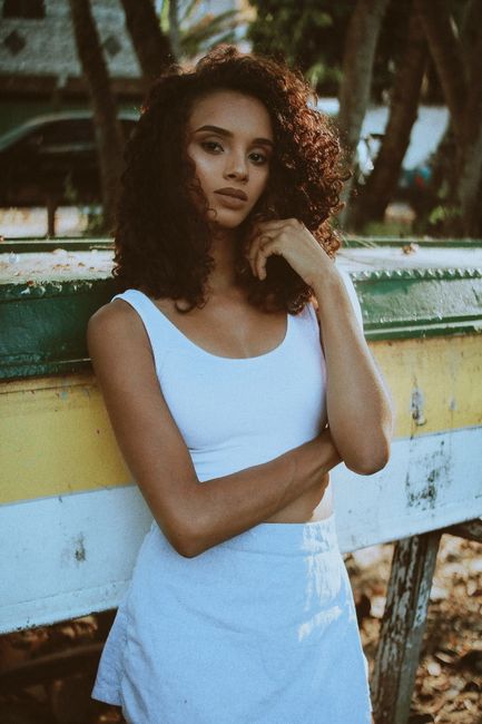 Stylish woman with curly hair poses by a boat in a tropical setting, exuding confidence.