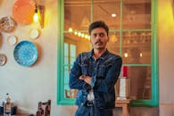 一个穿着牛仔夹克、留着小胡子的年轻人在室内摆姿势，背景是带有反光窗户和墙壁的装饰