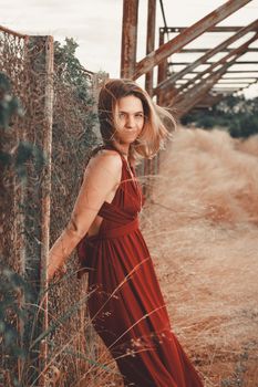 A blonde woman in a red dress leans against a rustic fence in an artistic outdoor setting.