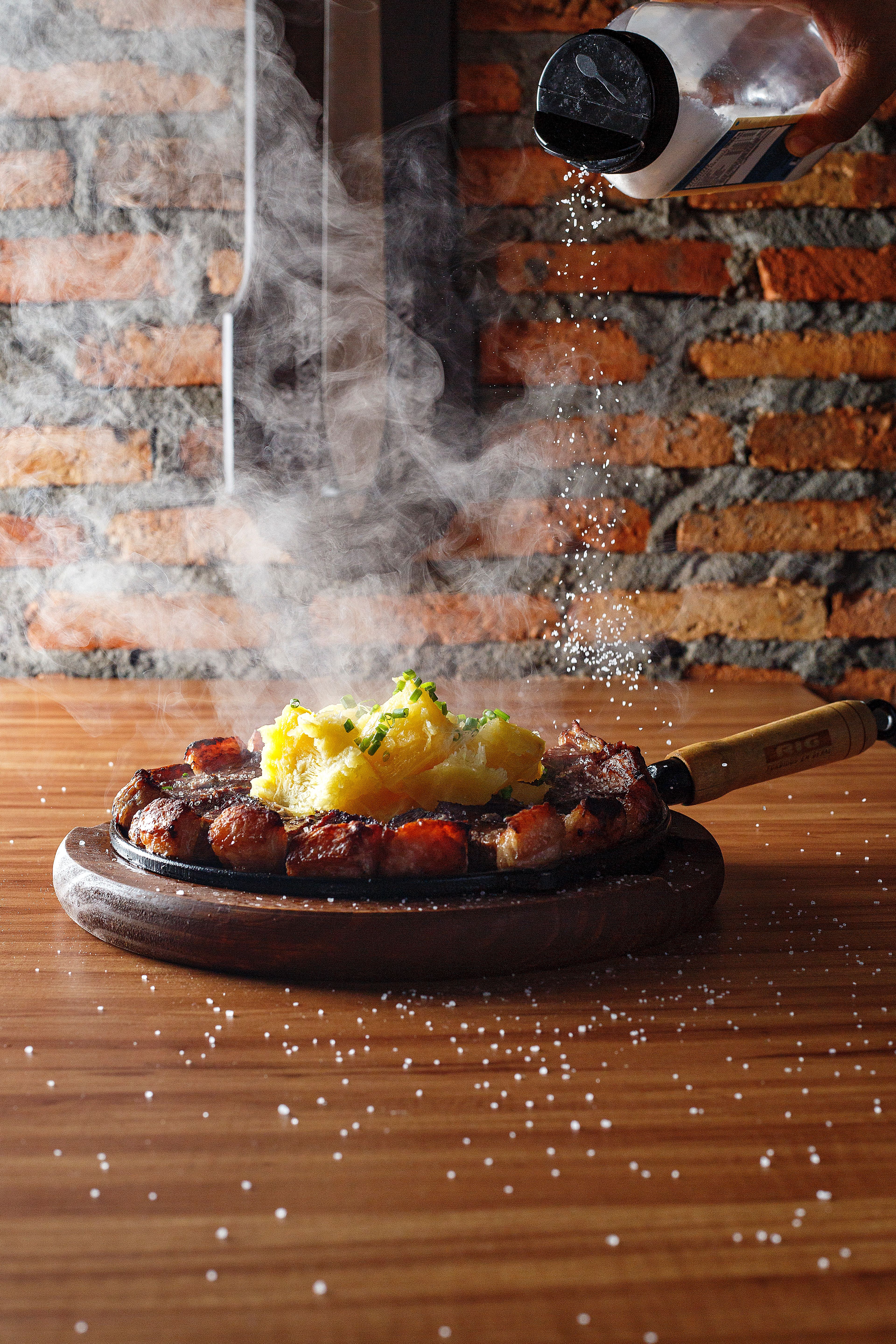 Close-up of a sizzling steak with mashed potatoes being seasoned with salt in a rustic setting.