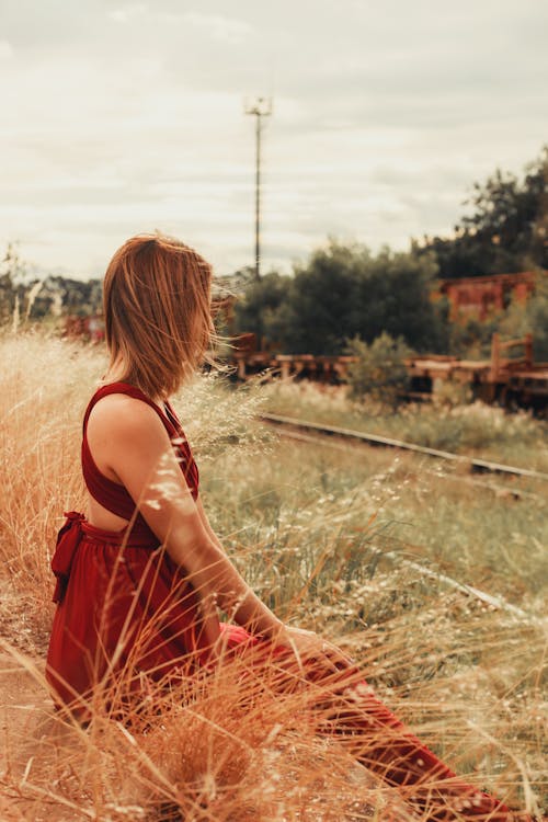 Woman in Red Gown Sitting in Field by Railway Tracks