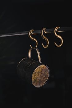 A vintage copper cup hanging on ornate copper hooks against a dark background.