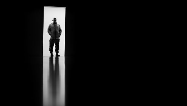 Silhouetted man in hoodie stands in doorway against black background. Dramatic lighting and contrast.