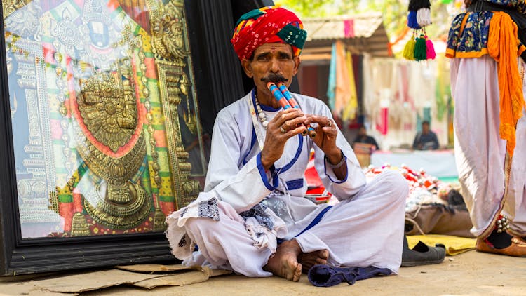 Old Man In Traditional Clothing Sitting On Ground Playing On Panpipe