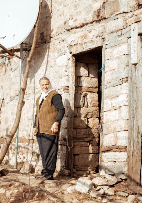 Elderly woman standing outside a rustic brick house in rural Tirana, Albania.