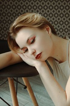 Stylish woman with artistic makeup resting on a chair indoors, evoking calmness.