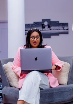Focused woman in pink blazer works on laptop seated on a cozy gray couch indoors.