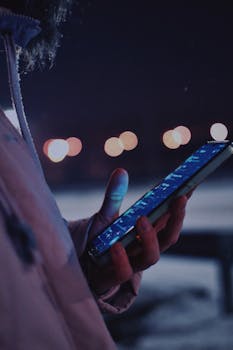 Close-up of hand holding smartphone illuminated by city lights during nighttime.