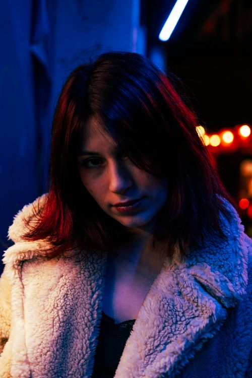 Free A woman in a fuzzy coat poses under neon lights at night, creating a moody urban vibe. Stock Photo