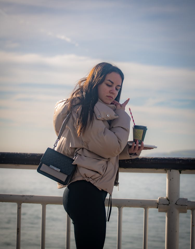 Woman Standing On Pier Overlooking Water Holding Phone And Coffee Cup In Hand