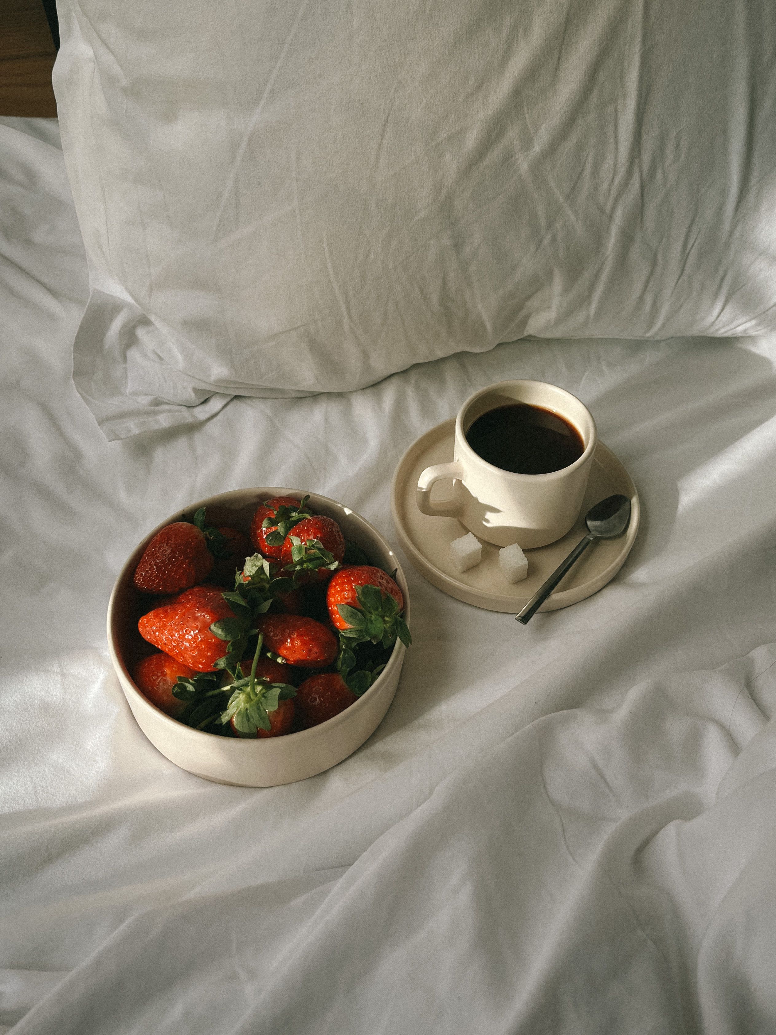 A bowl of fresh strawberries and a coffee cup on a bed with white sheets, capturing a cozy morning vibe.