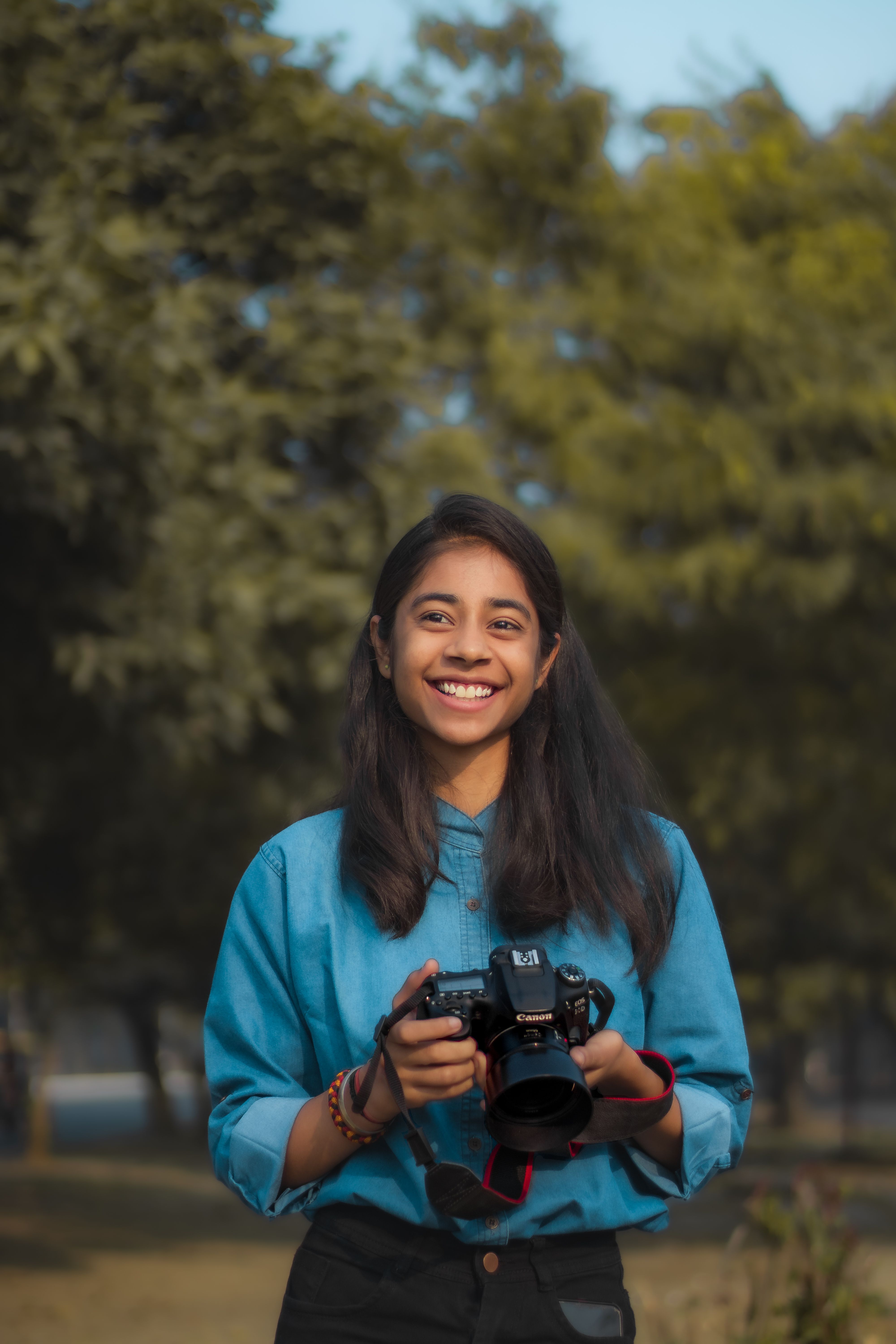 Teen girl smiling while holding a camera in a park setting during the day.