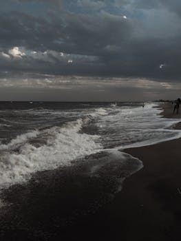 A dark and moody ocean scene at dusk, featuring crashing waves and a cloudy sky.