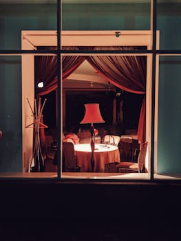 Warmly lit restaurant corner with an elegant lamp, table, and chairs viewed through a window.