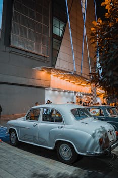 Retro Hindustan Ambassador car parked in urban nightlife setting with modern architecture background.