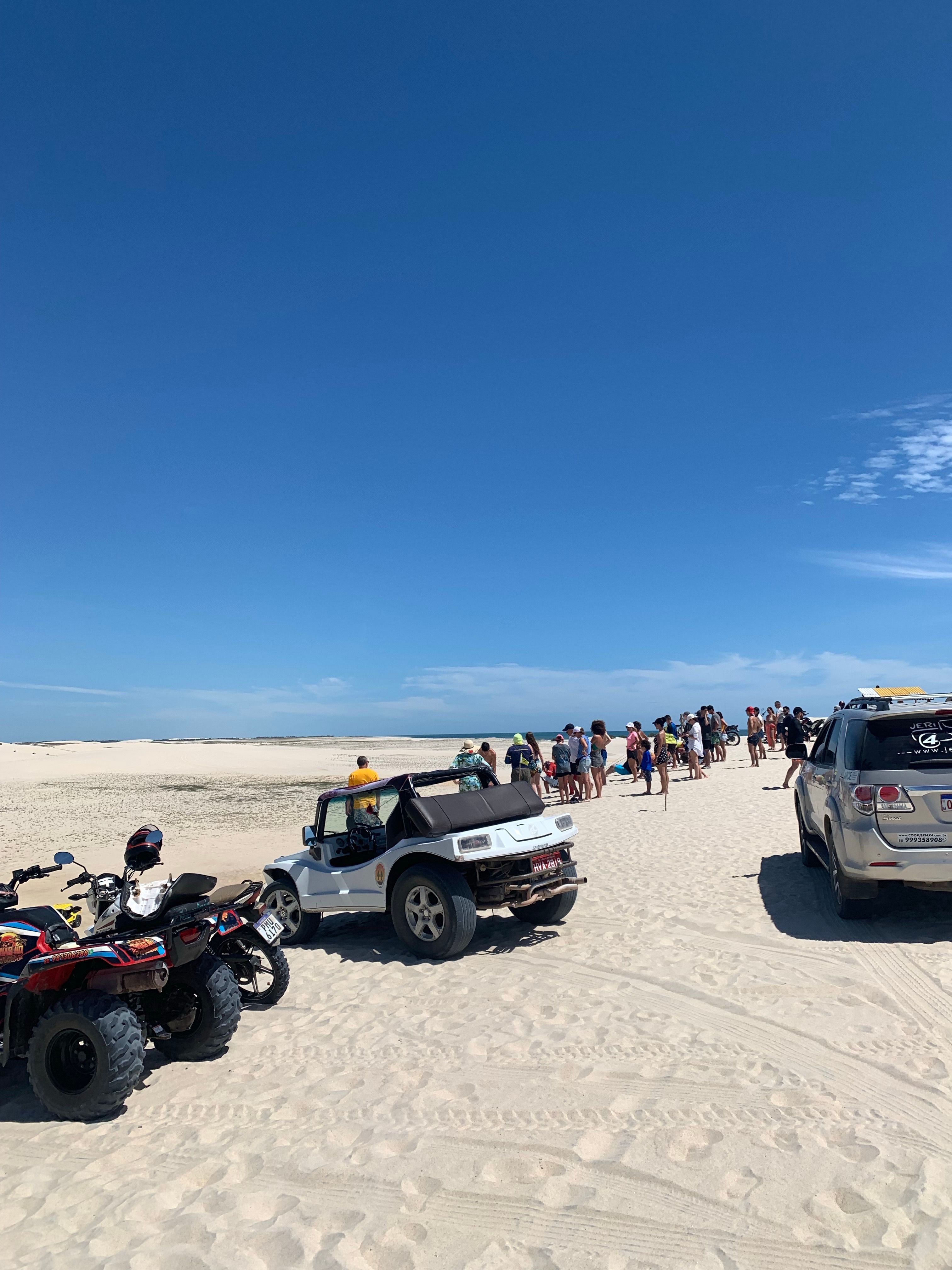 A group of off-road vehicles and people exploring a sandy desert under a clear blue sky.