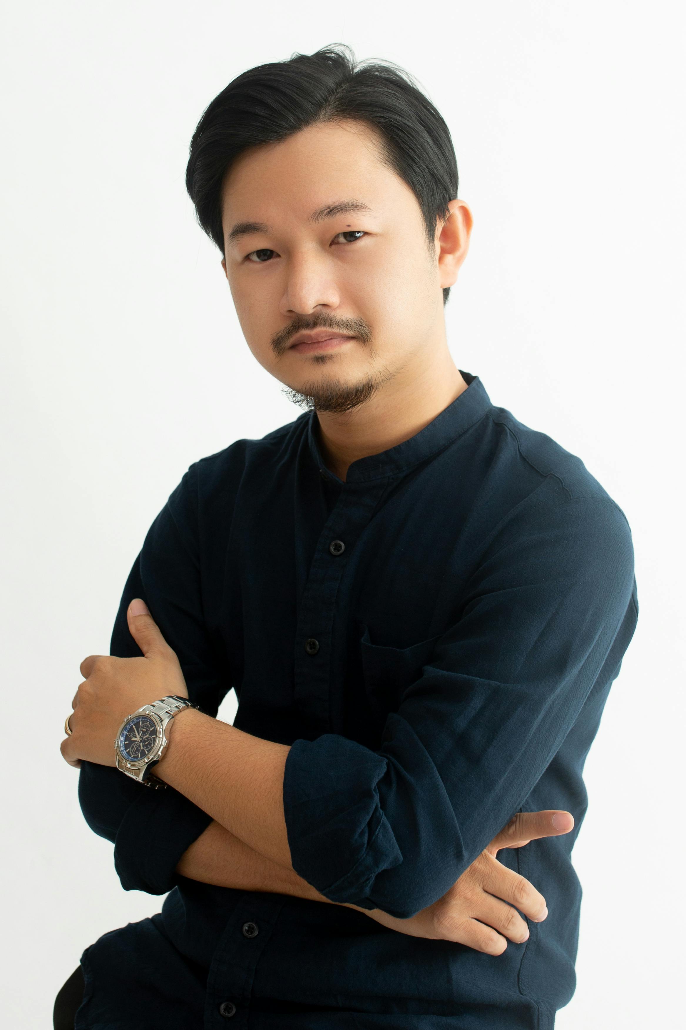 Studio portrait of a confident man with a mustache, wearing a dark shirt and a wristwatch.