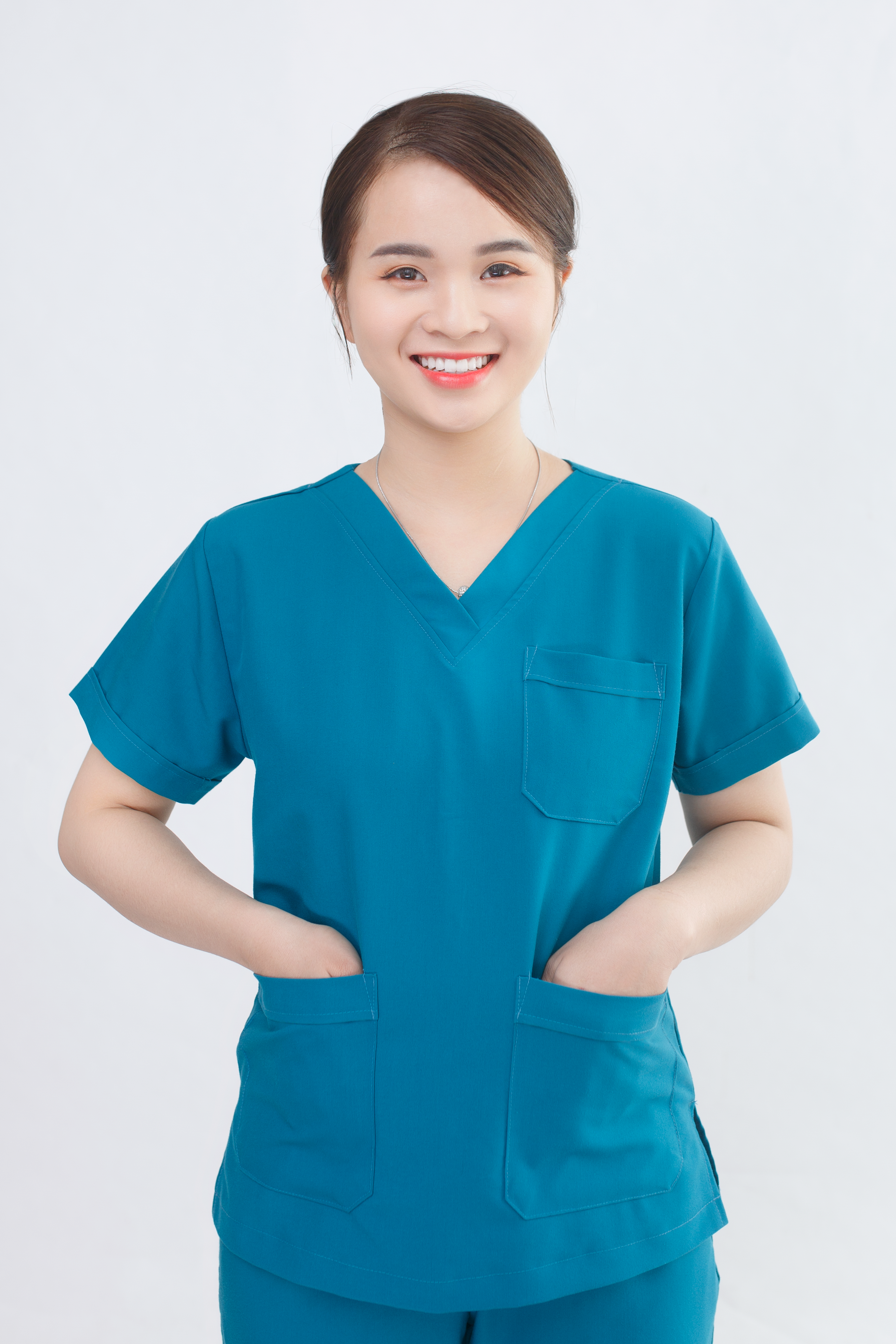 Smiling young nurse wearing blue scrubs, standing with hands in pockets against a white background.