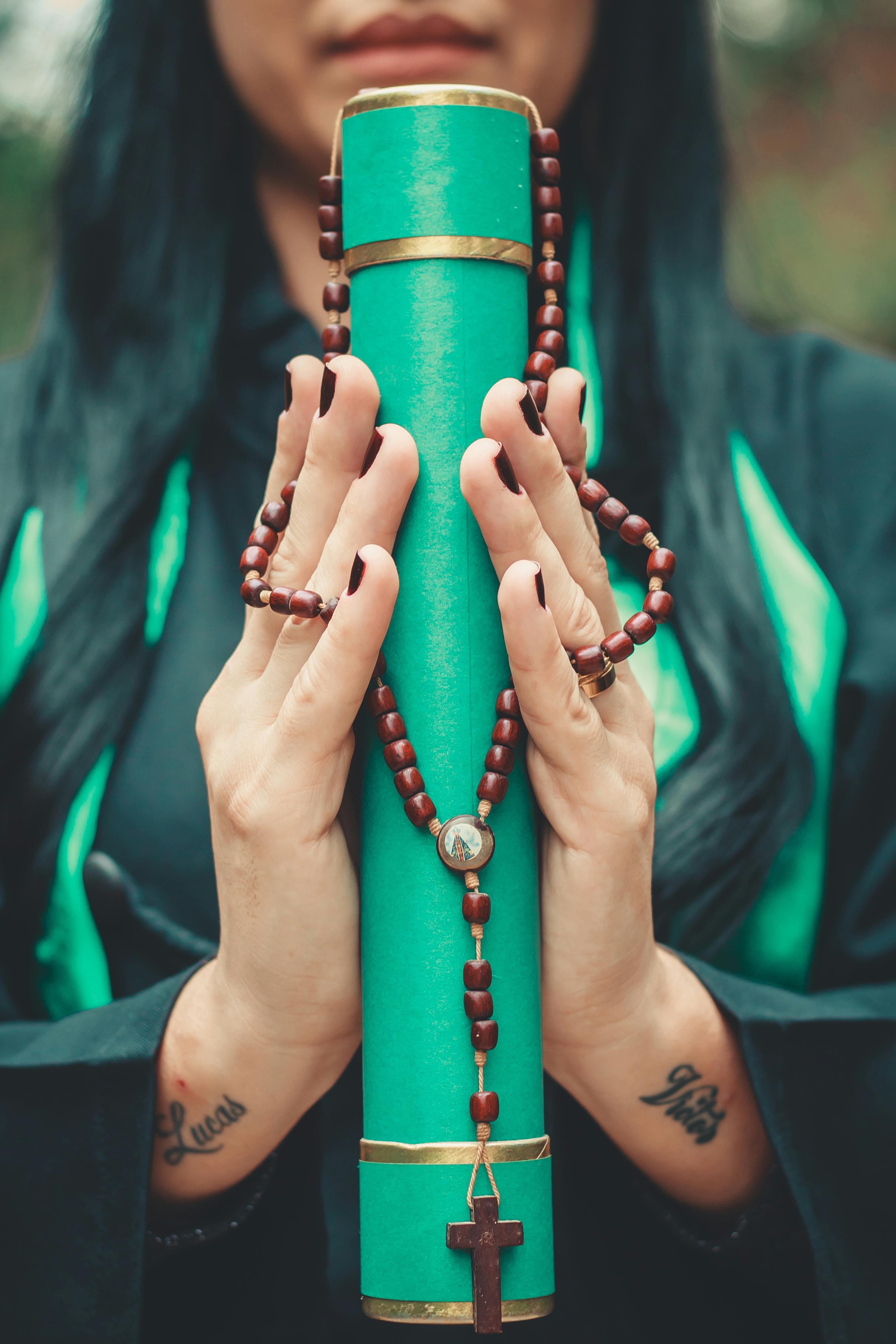 Close-up of woman's hands holding a graduation scroll adorned with a rosary, symbolizing faith and achievement.