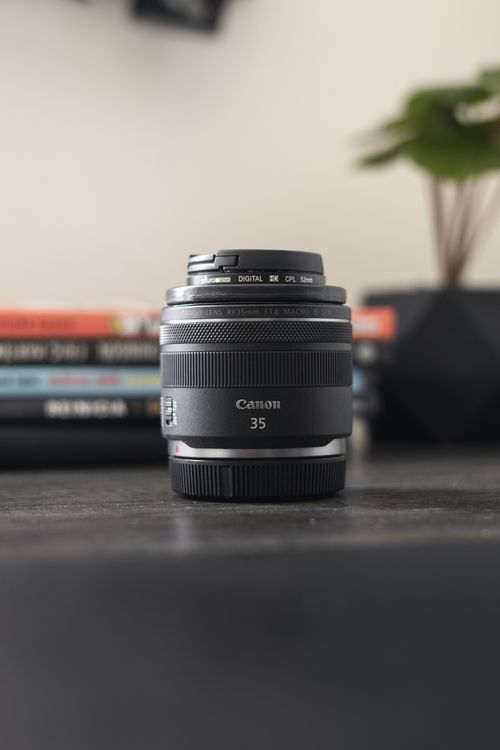 Free A Canon 35mm lens sits in focus on a desk with books and a plant in the background. Stock Photo
