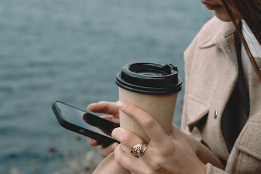 Close-up of a woman holding coffee and using smartphone near water. Stylish and serene.